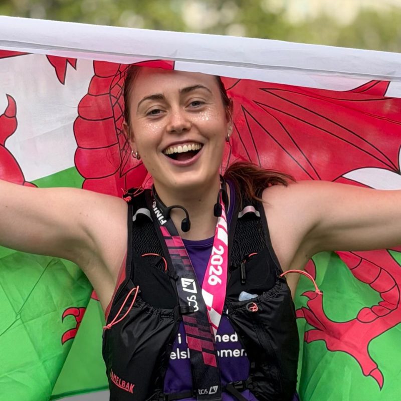 Woman smiling wearing a purple WWA running vest, holding a Welsh flag above her head.